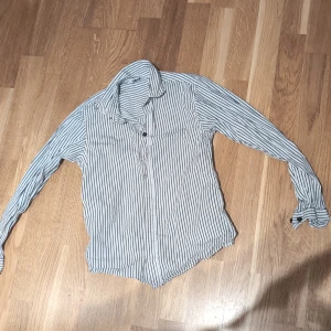 Vit och blå randig skjorta pojk strl 146 - Säljer en stilren vit skjorta med blå ränder. Skjortan har långa ärmar och knappar framtill. Perfekt för en avslappnad men ändå uppklädd look. Strl 146- pojk