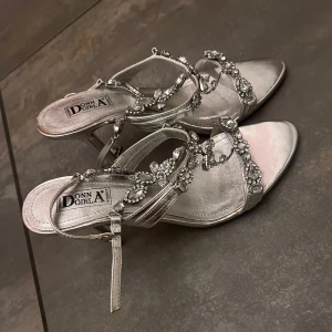 Klackar från Donna Girl - Eleganta silvriga klackar från Donna Girl med glittrande strassdetaljer. Sandalerna har en smal klack och justerbara remmar för perfekt passform. Perfekta för att ge en lyxig touch till din outfit.