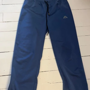 Blå mjukisbyxor från Kappa - Säljer ett par bekväma blå mjukisbyxor från Kappa med elastisk midja och snörning. Perfekta för en avslappnad stil. Byxorna har en liten logga på sidan och är i en loose passform. 💕Snöret är ej i orginal och det är resor, så du får välja om jag ska ta ur det innan jag postar eller om du vill ha kvar det.💕 Dem är köpta på second hand och redan då va lappar bortklippta💕