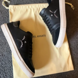 Svarta sneakers från Axel Arigato - Snygga svarta sneakers från Axel Arigato med fågelbroderi på sidan. Skorna har en vit sula och är tillverkade i skinn med snörning framtill. För en stilren look och en perfekt vår och sommar sko. 