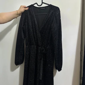 Svart glittrig klänning  - Snygg svart klänning med glittriga detaljer. Den har en v-ringad design och långa ärmar. Perfekt för en kväll ute eller en speciell tillställning. Klänningen har en knytning i midjan för en smickrande passform. Längden är över knäna. Priset kan förhandlas💕💗