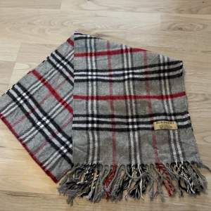 Grå rutig halsduk från Burberry - Säljer en klassisk grå rutig halsduk från Burberry i 100% lammull. Den har ett stilrent mönster med röda och svarta rutor samt fransar i ändarna. Perfekt för att hålla dig varm och snygg under kyliga dagar. Perfekt nu inför våren 