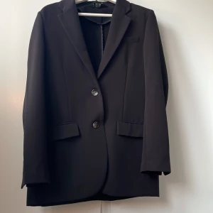 Svart kavaj med knappar - Elegant svart kavaj med två knappar framtill och klassiska slag. Perfekt för en stilren look. Kavajen har två fickor med lock och en bröstficka. Passar bra till både formella och semi-formella tillfällen.