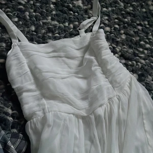 Vit klänning från Lindex - Säljer en söt vit klänning från Lindex med smala axelband och en smockad rygg. Klänningen har en luftig och flödande design, perfekt för varma dagar. Den är tillverkad i ett lätt och bekvämt material.