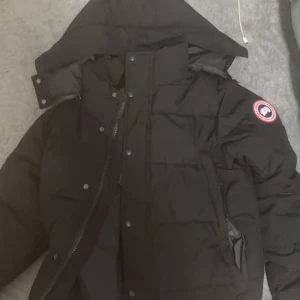Svart dunjacka från Canada Goose - Säljer en svart dunjacka från Canada Goose med huva och dragkedja. Jackan har en broderad logotyp på ärmen och är perfekt för kalla dagar. Den är både stilren och funktionell med flera praktiska fickor.