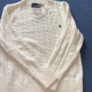 Vit stickad tröja från Polo Ralph Lauren - Säljer en stilren vit stickad tröja från Polo Ralph Lauren med kabelstickat mönster och broderad logga på bröstet. Tröjan har långa ärmar och ribbade muddar vid ärmslut och nederkant. Perfekt för en klassisk look. Fick denna i present men passar inte riktikgt min stil