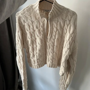 Beige kabelstickad kofta från NA-KD - Säljer en snygg beige/vit/cremefärgad kabelstickad kofta från NA-KD med ett flätat mönster. Koftan har en hög krage och är croppad, vilket ger den en modern look. Perfekt för kyligare dagar!