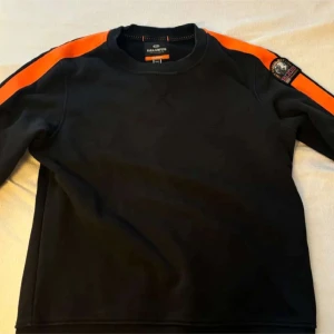 Svart tröja med orange detaljer från Parajumpers - Snygg svart tröja från Parajumpers med orangea ränder längs ärmarna. Tröjan har en rund hals och en patch på ärmen. Perfekt för en sportig look. Skick 10/10 har aldrig används 