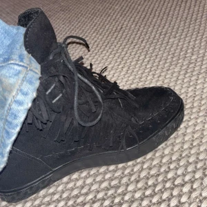 Svarta mocka sneakers med fransar - Snygga svarta sneakers i mocka med fransdetaljer och snörning. Perfekta för en trendig look med en twist. Köpte på Plick men det passade inte💔klacken är inte jätte hög. Vet inte märke, skriver de hon jag köpte de av sa