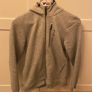 Grå hoodie från Sail Racing - Storlek M - Säljer en stilren grå hoodie från Sail Racing med dragkedja och huva. Tröjan har långa ärmar och en broderad logga på bröstet. 