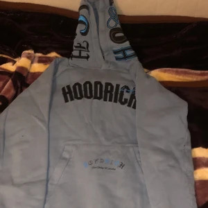 Ljusblå hoodie från Hoodrich - Säljer en ljusblå hoodie från Hoodrich med tryck både fram och bak. Den har en stor ficka framtill och en mönstrad huva med textdetaljer. Perfekt för en avslappnad stil.
