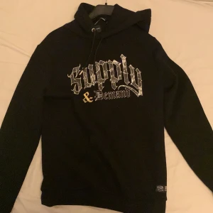 Svart hoodie från Supply & Demand - Säljer en svart hoodie från Supply & Demand med tryck på framsidan. Hoodien har en klassisk design med justerbar huva och ribbade muddar. Perfekt för en avslappnad stil.