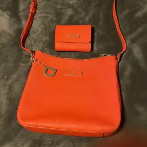 Snygg orange axelväska från DKNY med matchande plånbok. Väskan har en justerbar axelrem och dragkedja upptill. Plånboken har flera fack och en knappstängning. Perfekt för att hålla ordning på dina saker med stil. Man får båda för 700🧡