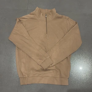 Beige tröja från Elitzia Junior - Säljer en beige tröja från Elitzia Junior med en halv zip vid halsen. Tröjan har långa ärmar och ribbade muddar vid ärmslut och nederkant. Perfekt för kyligare dagar. 