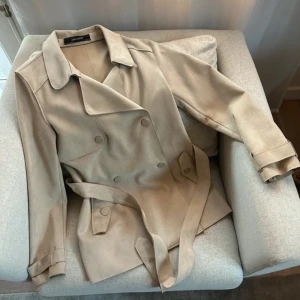 Beige trenchcoat från Zara - Snygg beige trenchcoat från Zara Basic med klassisk dubbelknäppning och bälte i midjan. Jackan har långa ärmar och en stilren design som passar perfekt för vår och höst. 