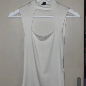 Vit ärmlös topp med hög krage - Snygg vit ärmlös topp med hög krage och stilren design. Toppen har en unik detalj med en öppning framtill som ger en modern touch. Perfekt för att styla med olika outfits.