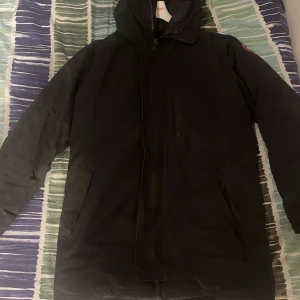 Svart dunjacka från Canada Goose - Säljer en svart dunjacka från Canada Goose med huva och dragkedja. Jackan har en klassisk design med långa ärmar och är perfekt för kalla dagar. Märkeslogga synlig på ärmen och dragkedjan.