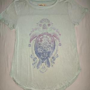 Mintgrön t-shirt med sköldpaddsmotiv från Hollister - Säljer en mintgrön t-shirt från Hollister med ett stort, färgglatt sköldpaddsmotiv i lila och blått på framsidan. T-shirten har en rund halsringning och korta ärmar. Perfekt för en avslappnad stil!