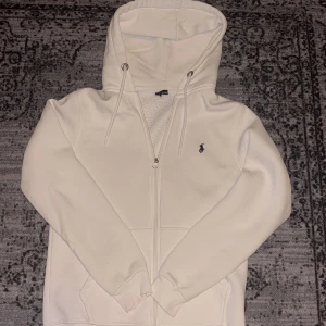Vit hoodie från Ralph Lauren - Säljer en stilren vit hoodie från Ralph Lauren med dragkedja och det klassiska broderade logotypen på bröstet. Tröjan har långa ärmar och en praktisk huva med snörning. Perfekt för en avslappnad look.