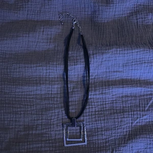Svart halsband med fyrkantig hängsmycke - Elegant svart halsband med ett fyrkantigt hängsmycke. Hängsmycket har en glittrig kant som ger en stilren look. Halsbandet har en justerbar kedja för perfekt passform.