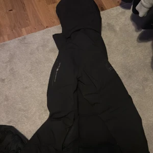 Svart dunjacka från Sail racing - Säljer en svart dunjacka från Peak Performance. Jackan har en huva och dragkedja framtill. Perfekt för kalla dagar med sin varma och skyddande design.