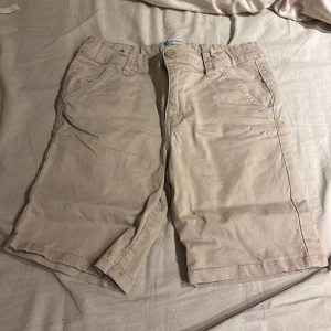 Beige shorts från Shill - Säljer ett par beige shorts från Shill. De har en klassisk design med knappar och fickor både fram och bak. Perfekta för en avslappnad stil.