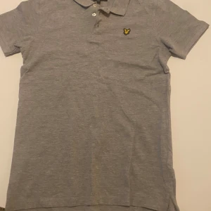 Grå pikétröja från Lyle & Scott - Säljer en stilren grå pikétröja från Lyle & Scott med korta ärmar och klassisk krage. Tröjan har en broderad logotyp i gult på bröstet och knappar framtill. Perfekt för en avslappnad men ändå snygg look. Hör av er vid funderingar!