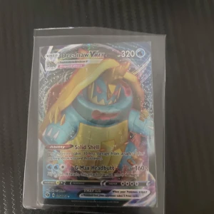 Drednaw VMAX Pokémonkort - Säljer ett Drednaw VMAX Pokémonkort med 320 HP. Kortet har förmågan 'Solid Shell' och attacken 'G-Max Headbutt' med 160+ skada. Perfekt för samlare eller spelare som vill förstärka sin kortlek.