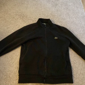 Svart tröja från Lacoste - Säljer en stilren svart tröja från Lacoste med dragkedja och det klassiska krokodilmärket på bröstet. Jackan har långa ärmar och en normal passform. Använd men inte sliten