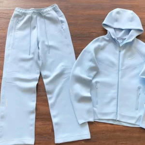 Ljusblå hoodie och byxor från Nike - Säljer en ljusblå hoodie och matchande byxor från Nike. Hoodien har dragkedja och en justerbar huva, medan byxorna har elastisk midja med dragsko. Båda plaggen har diskreta Nike-loggor och är tillverkade i ett mjukt syntetmaterial.
