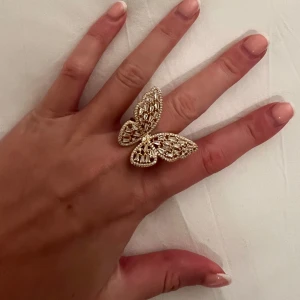 Guldfärgad ring med fjärilsdesign - Elegant guldfärgad ring med en stor fjärilsdesign. Fjärilen är dekorerad med glittrande stenar som ger en lyxig känsla. Perfekt för att ge en extra touch till din outfit.