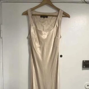 Champagnefärgad sidenklänning - Elegant champagnefärgad klänning i siden med en smickrande passform. Klänningen är ärmlös och har en mjuk, glansig yta som ger en lyxig känsla. Perfekt för speciella tillfällen.