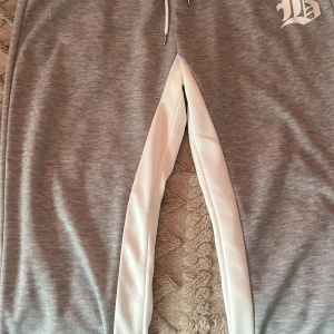 Grå mjukisbyxor med vit detalj - Snygga grå mjukisbyxor med vita sidopaneler och en tryckt bokstavsdetalj. Byxorna har snörning i midjan för justerbar passform. Perfekta för en avslappnad stil. Byxorna sitter bra vid midjan ner mot låret sedan sitter löst vid foten alltså ändan av byxan.Pris kan diskuteras