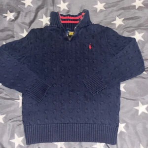 Mörkblå halvzip Ralph Lauren (barn) - Säljer en snygg mörkblå stickad tröja från Ralph Lauren med dragkedja och röd logga på bröstet. Tröjan har en ribbad krage med röd och blå randig insida, samt långa ärmar och ribbade muddar. 