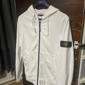 Vit hoodie från Stone Island - Säljer en stilren vit hoodie från Stone Island med dragkedja och huva. Tröjan har en ikonisk logga på ärmen och praktiska fickor framtill. Perfekt för en avslappnad stil.