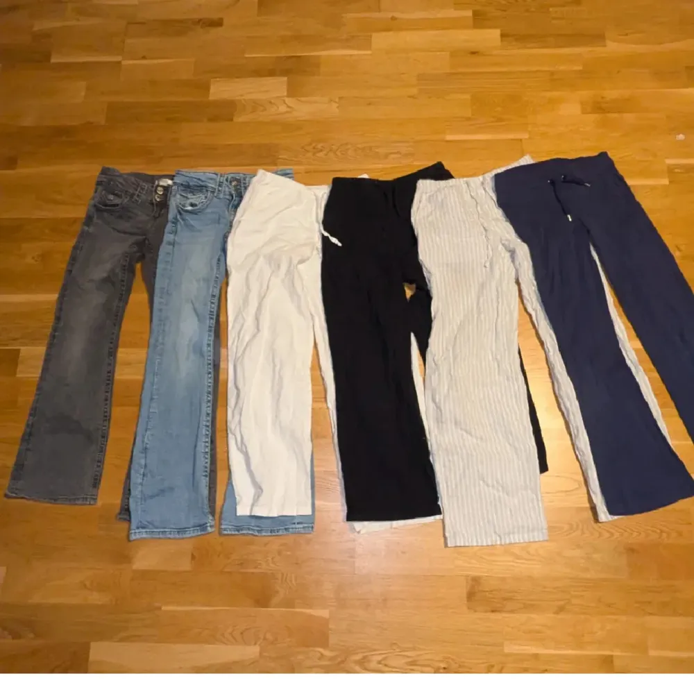 En samling av sex olika byxor i varierande färger och stilar. Inkluderar jeans i grått och ljusblått, vita och svarta byxor samt randiga och mörkblå byxor. Perfekt för att variera garderoben med olika passformer och material. En liten fläck befinner sig på den vita linnebyxan.. Farkut & Housut.
