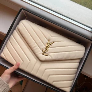 Beige quiltad axelväska från Saint Laurent - Säljer en elegant beige axelväska från Saint Laurent med quiltad design och guldfärgade kedjedetaljer. Väskan har en ikonisk logotyp framtill och ett rymligt inre med dragkedjefack. Perfekt för att ge en lyxig touch till din outfit. Osäker på äktheten då jag har fått den. Jättefin väska. Har endast haft den i garderoben så är inte använd<33 färgen är som mest lik som på första bilden 