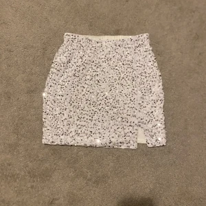 Vit paljettkjol - Snygg vit kortkjol täckt med glittriga paljetter. Kjolen har en liten slits framtill och en elastisk midja för bekväm passform. Perfekt för att lysa upp vilken outfit som helst! M men sitter som S