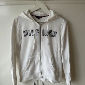 Vit hoodie från Tommy Hilfiger - Säljer en stilren vit hoodie från Tommy Hilfiger med dragkedja och huva. Den har märkets namn broderat på framsidan i svart. Perfekt för en avslappnad look.