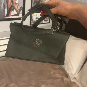 Grön handväska med metallogo - Snygg grön handväska med en stilren metallogo på framsidan. Väskan har ett lock med asymmetrisk design och ett bekvämt handtag. 