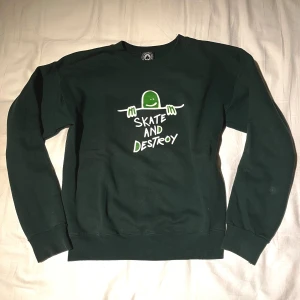 Thrasher sweatshirt  - Säljer en grön sweatshirt med ett coolt tryck 'Skate and Destroy' på framsidan. Tröjan har långa ärmar och är perfekt för en avslappnad stil. Den är gjord i ett mjukt material. Den har en liten fläck på under armen. 