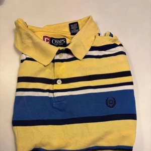 Randig pikétröja från Chaps - Säljer en färgglad randig pikétröja från Chaps i gult, blått och svart. Tröjan har en klassisk krage med knappar och ett broderat emblem på bröstet. Perfekt för en avslappnad stil. 