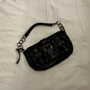 Svart axelväska med kedjedetaljer - Snygg svart liten axelväska i skinn med coola kedjedetaljer på axelremmen. Väskan har två framfickor med spännen och en dragkedja upptill🥀