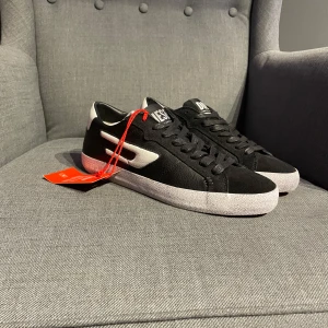 Svarta sneakers från Diesel - Snygga svarta sneakers från Diesel med vit sula och detaljer. Skorna har snörning och en stilren design med märkets logga på plösen. Perfekta för en casual look. Helt nya aldrig använda. Ny pris 1300