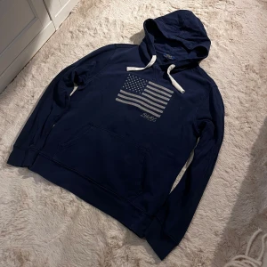 Mörkblå hoodie från Polo Ralph Lauren - Säljer en snygg mörkblå hoodie från Polo Ralph Lauren med en stor amerikansk flagga på bröstet. Hoodien har en klassisk känguruficka och vita dragsnören vid huvan. Perfekt för en avslappnad stil.