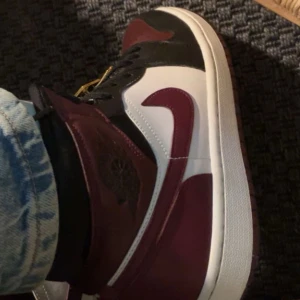 Nike Jordan 1 'Dark Beetroot' Mid - Snygga Nike Jordan 1 'Dark Beetroot' Mid i en cool färgkombination av vinröd, svart och vit. Skorna har den klassiska Air Jordan-designen med snörning och en hög siluett. Perfekta för dig som vill ha en stilren och sportig look.