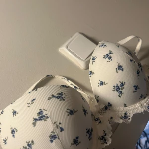 Vit blommig bh med spetsdetaljer - Söt vit bh med blå blommönster och spetsdetaljer längs kanterna. Den har en liten rosett framtill och justerbara axelband för bästa passform. Perfekt för en romantisk look. HELT NY BARA BORT TAGNA LAPPAR!!