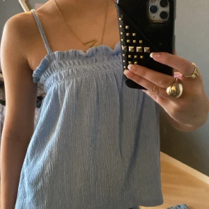 Ljusblå topp från Gina Tricot - Söt ljusblå topp från Gina Tricot med smala axelband och en rynkad design. Perfekt för varma dagar med sin luftiga och bekväma passform. Toppen har en fin volangdetalj vid halsringningen som ger en extra touch av stil.