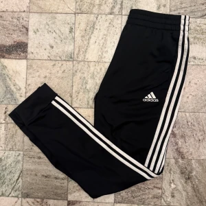 Svarta träningsbyxor från Adidas - Snygga svarta träningsbyxor från Adidas med de klassiska tre vita ränderna längs sidorna. Byxorna har en elastisk midja och är perfekta för sport och träning. Adidas-loggan är tryckt på framsidan.