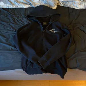 Svart hoodie från Peak Performance - Säljer en svart hoodie från Peak Performance med dragkedja och huva. Perfekt för kyliga dagar och har en stilren design med logotyp på bröstet. Passar både till vardags och träning.